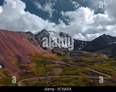 Panoramablick auf eine kurvenreiche Straße, die sich entlang leuchtender roter und grüner alpiner Hänge in Richtung einer massiven, schneebedeckten Bergkette schlängelt Stockfoto