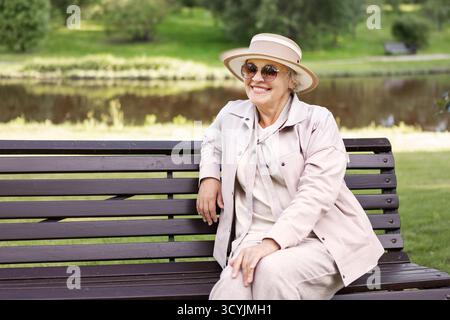 Schöne Seniorin, die sich am sonnigen Tag auf der Parkbank entspannt, lächelt und im Ruhestand friedliche Momente im Freien genießt, einen stilvollen Hut trägt und Stockfoto