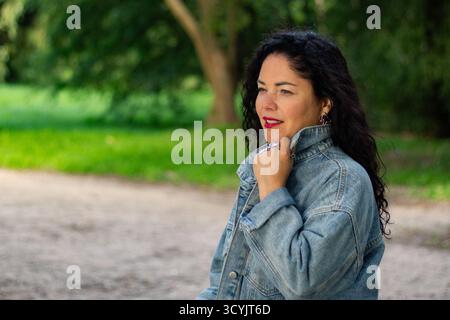 Porträt einer selbstbewussten lockigen Frau in der Natur. Nahaufnahme-Porträt einer dunkelhaarigen Frau mit voluminösen Locken, die selbstbewusst vor verschwommenem Grün lächelt Stockfoto