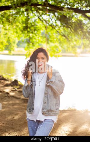Porträt einer selbstbewussten lockigen Frau in der Natur. Nahaufnahme-Porträt einer dunkelhaarigen Frau mit voluminösen Locken, die selbstbewusst vor verschwommenem Grün lächelt Stockfoto