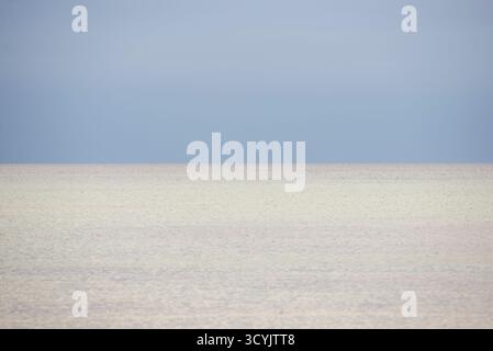 Eine ruhige Küstenszene mit ruhigem, hellem Wasser, das sich in einen sanften blauen Himmel verwandelt. Der Horizont ist kaum sichtbar, was eine minimalistische Komposition mit sich bringt Stockfoto