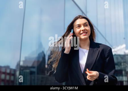 Eine junge Frau steht draußen, lächelt und verwickelt sich in ein Telefongespräch. Sie trägt ein modernes Outfit, während sich das Sonnenlicht in der Nähe von G reflektiert Stockfoto