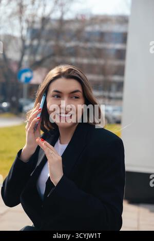 Eine junge Frau mit langen Haaren lächelt, während sie draußen ein Telefongespräch führt. Sie wirkt fröhlich und trägt legere Kleidung in einer urbanen Umgebung mit bl Stockfoto