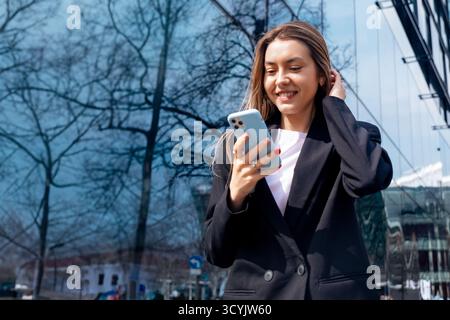 Eine Frau interagiert lässig mit ihrem Smartphone vor einem eleganten Glasgebäude und lächelt, während sie an einem hellen, sonnigen Tag ihre Zeit im Freien genießt. Stockfoto