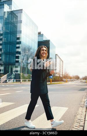 Eine junge Frau schlendert selbstbewusst über einen Stadtübergang, mit ihrem Smartphone, umgeben von eleganten Glasgebäuden an einem sonnigen Tag. Stockfoto