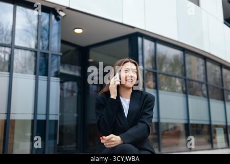 Eine Frau in einem stilvollen schwarzen Anzug sitzt auf einer Bank, lächelt, während sie mit ihrem Smartphone vor einer modernen Geschäftsstruktur auf einer sonnigen Sonne spricht Stockfoto