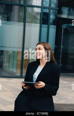 Eine Frau in einem schwarzen Blazer sitzt bequem im Freien und lächelt, während sie mit einem Tablet interagiert. Sie steht an einem sonnigen Tag vor einem eleganten Büro Stockfoto