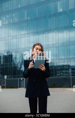 Eine Frau in einem maßgeschneiderten schwarzen Anzug steht draußen und blickt auf ihr Tablet, während sie von einem eleganten Bürogebäude aus Glas umgeben ist. Sie scheint konzentriert in ihr zu sein Stockfoto