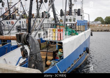 Das Heck eines blau-weißen Fischtrawlers dockte in einem belebten Hafen an, beladen mit Fischernetzen, Kisten und Seilen, mit anderen Schiffen im Hintergrund. Stockfoto