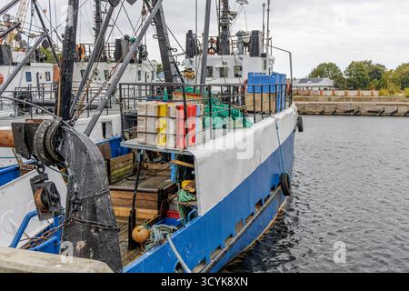 Das Heck eines blau-weißen Fischtrawlers dockte in einem belebten Hafen an, beladen mit Fischernetzen, Kisten und Seilen, mit anderen Schiffen im Hintergrund. Stockfoto
