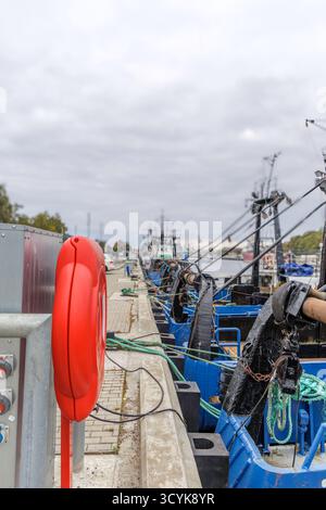 Vertikale Nahaufnahme eines orangefarbenen Rettungsschirms und elektrischer Versorgungskästen auf einem Betondock, mit einer Linie blauer und weißer Fischfänger, die dahinter verankert sind. Stockfoto