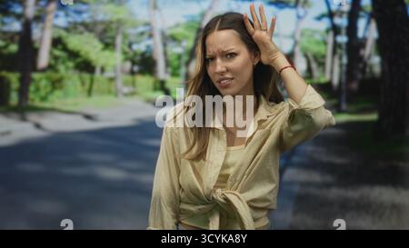 Junge Frau, die Hand an Ohr auf der Straße neben dem grünen Parkweg hält, Hemd gebunden und schielender Ausdruck; Verwirrung. Stockfoto