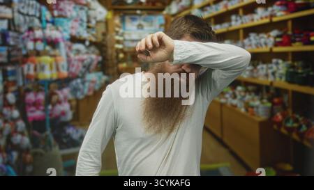 Bärtiger Mann trocknet Schweiß auf der Stirn in einem mit Holzregalen und farbenfrohen Waren gefüllten Innengang; Müdigkeit. Stockfoto