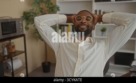 Mann mit Brille und Zahnspange, Hände hinter dem Kopf lächelnd am Schreibtisch im Büro, mit weißem Hemd; Karrierezufriedenheit. Stockfoto