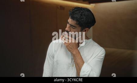 Mann mit Hand am Kinn und nachdenklichem Ausdruck in weißem Hemd in einem minimalistischen, braunen Studio-Raum; Zweifel an der Kontemplation. Stockfoto