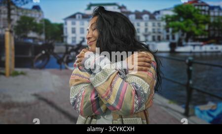 Frau umarmt ihre Schultern auf der Straße am amsterdamer Kanal mit geschlossenen Augen und einem leichten Lächeln; Selbstfürsorge-Gelassenheit. Stockfoto