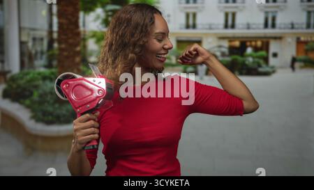 Frau, die Packband in der Hand hält und lächelt, während sie in einer Fußgängerzone mit Gebäudefassade Bizeps bicelt; Zuversicht. Stockfoto