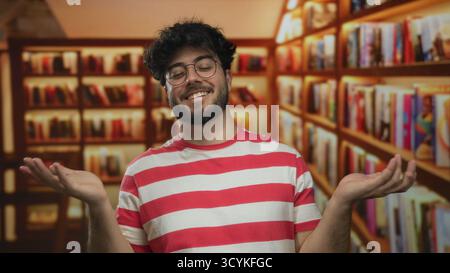 Junger bärtiger hispanischer Mann mit Brille und gestreiftem Hemd steht selbstbewusst in einer Bibliothek, umgeben von Regalen mit bunten Büchern, die eine intel-information erschaffen Stockfoto