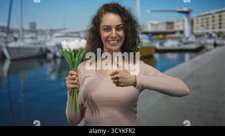 Junge hispanische Frau, die weiße Tulpen in der linken Hand hält und mit der rechten Hand auf einer Straße neben Booten und Yachthafen Daumen nach oben gibt; Glück. Stockfoto