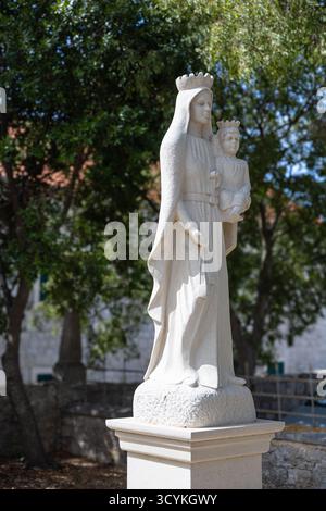 Eine heitere Statue aus weißem Stein der Jungfrau Maria, die das Kind in der Hand hält, gekrönt auf einem Podest in einem sonnendurchfluteten Garten steht. Perfekt für religiöse, Gedenkstätten oder Stockfoto