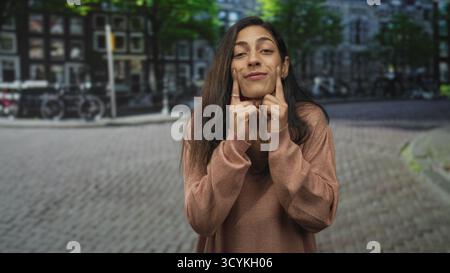 Frau, die die Wangen mit Zeigefingern auf einer Kopfsteinpflasterstraße auf einem Stadtplatz drückt, mit Blick auf die Kamera; verspielter Ausdruck. Stockfoto