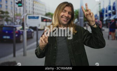 Mann mit grüner Cordjacke hebt beide Hände und macht Friedenszeichen und lächelt breit auf der überfüllten Stadtstraße; Glück. Stockfoto