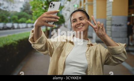 Hispanische Frau in einem lässigen Hemd, das Smartphone auf Armlänge hält, ein Friedensschild macht und lächelt auf einer Stadtstraße in der Nähe eines Steingebäudes; verspieltes c Stockfoto