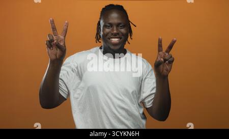 Junger afroamerikanischer Mann in weißem T-Shirt mit doppelten Friedenszeichen mit beiden Händen im Studio mit orangefarbenem Hintergrund; Glück Frieden Positivität. Stockfoto