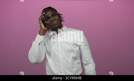 Mann, der mit der Hand das Ohr berührt und im Studio eine Brille vor rosa Hintergrund trägt; nachdenkliche Konzentration. Stockfoto