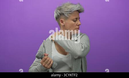 Frau mit kurzen Haaren in grauem Hoodie, die Hantel hält und den Hals im lila Studio umklammert; Unwohlsein. Stockfoto
