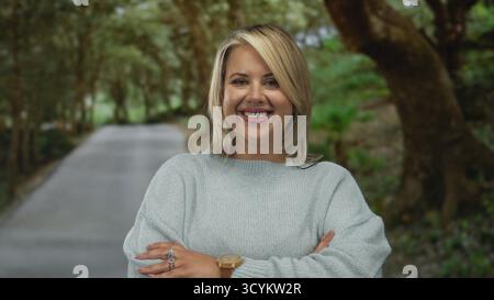 Junge blonde Frau in blauem Pullover lächelt mit überkreuzten Armen auf einem gepflasterten Weg im grünen Wald; Leugnung. Stockfoto