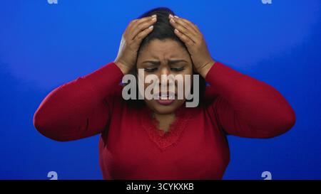 Frau, die Frustration vor einem leuchtend blauen Hintergrund ausdrückt, Hände auf dem Kopf trägt, ein rotes Hemd trägt, Emotionen und Isolation in einem Studio-Set vermittelt Stockfoto