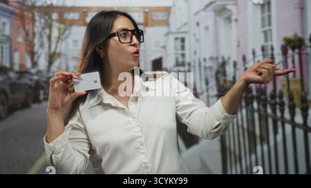 Junge hispanische Frau, die eine Brille trägt und Kreditkartenpunkte mit offenem Mund auf der Straße hält; überraschende finanzielle Dringlichkeit. Stockfoto