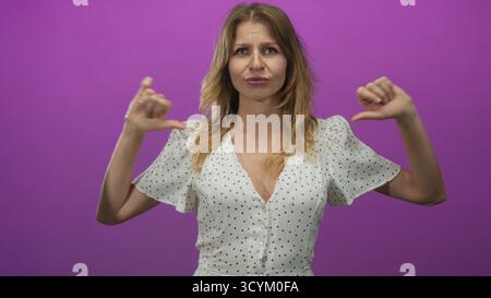 Frau, die beide Daumen auf die Brust zeigt im Studio mit violettem Hintergrund, weißes Kleid mit Punktmuster und geschnürten Lippen; Skepsis Selbstidentität. Stockfoto