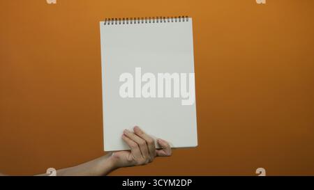 Mann mit leerem Notizblock mit weißen Seiten vor orangefarbenem Hintergrund, der kreativen Raum und isolierte Ideen in einem minimalistischen Ambiente betont. Stockfoto