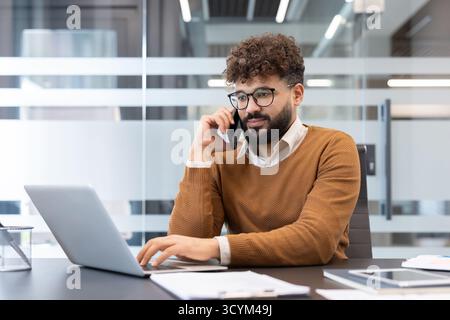 Junger bärtiger Unternehmer mit Brille, der sich an einem Telefongespräch beteiligt, während er aktiv auf einem Laptop tippt und mehrere Aufgaben von seinem modernen Schreibtisch aus verwaltet Stockfoto