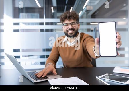 Der lächelnde junge Geschäftsmann im modernen Büro hält ein Smartphone mit leerem weißen Bildschirm für App-Mockup, während er am Laptop arbeitet, auf mobile Dienste oder digitale Produkte zeigt und präsentiert Stockfoto