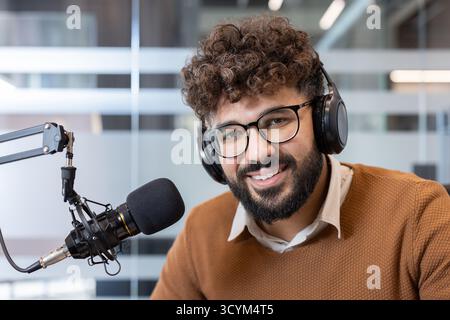 Junger Mann mit lockigen Haaren und Brille lächelt während er in einem modernen Studio einen Podcast aufnimmt, Kopfhörer trägt und in ein Mikrofon für Online-Audioinhalte spricht Stockfoto