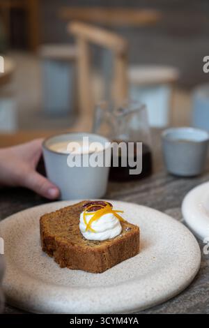 Ein Stück Karottenkuchen mit Frischkäse-Zuckerguss und Orangenschale wird auf einer Keramikplatte in einem gemütlichen Café serviert Stockfoto