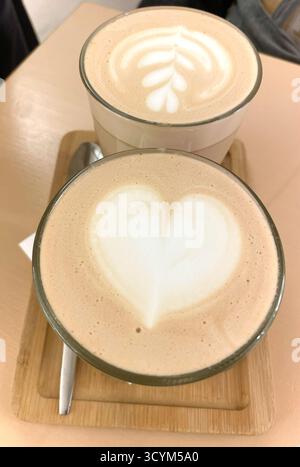 Zwei wunderschön gefertigte Kaffeegetränke mit komplizierten Latte Art Designs, eines mit Herz und das andere mit Blattmuster, präsentiert auf einem Holztablett Stockfoto