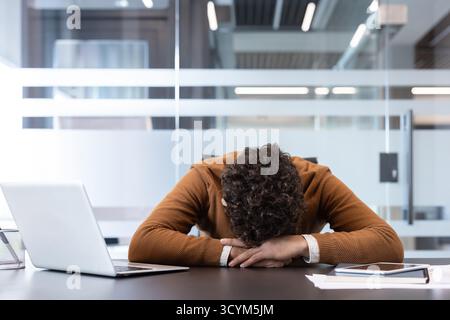 Gestresster Mann, der beruflich Burnout und Überlastung erlebt, seinen Kopf mit einem Laptop auf einem Schreibtisch ablegt und die Herausforderungen der psychischen Gesundheit in einer modernen Büroumgebung reflektiert Stockfoto