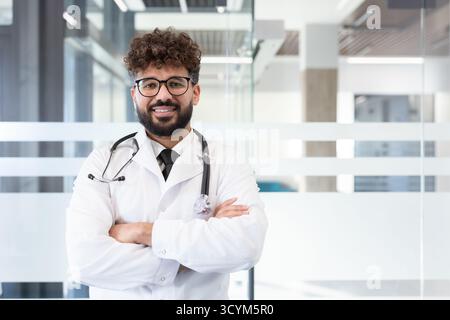 Junger männlicher Arzt mit weißem Labormantel und Stethoskop, freundlich lächelnd mit überkreuzten Armen, selbstbewusst in einem modernen Krankenhaus oder einer Klinik Stockfoto
