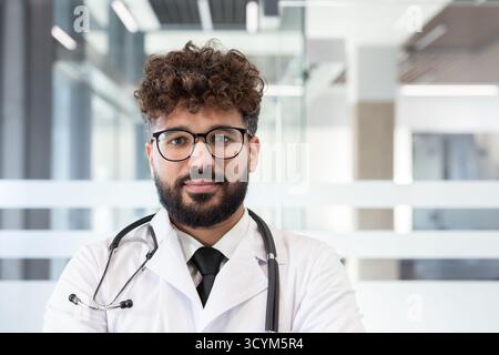 Junger männlicher Arzt mit Bart und lockigem Haar, Brille, Stethoskop und weißem Fell, selbstbewusst in einer modernen medizinischen Klinik oder Krankenhausflur Stockfoto