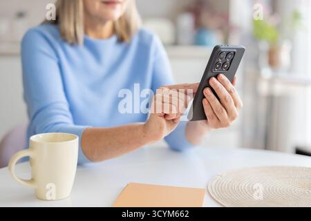 Eine Seniorin sitzt an einem weißen Tisch, hält ein schwarzes Smartphone, berührt den Bildschirm mit dem Finger und beschäftigt sich mit digitalen Inhalten und Technologien in ihrer modernen Wohnumgebung Stockfoto