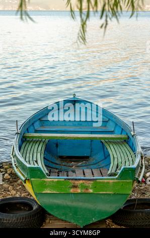 Altes grünes und blaues hölzernes Ruderboot am ruhigen Seeufer mit sanften Reflexen auf der friedlichen Wasseroberfläche Stockfoto