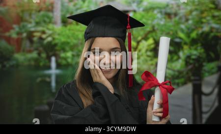 Teenager-Mädchen mit Mütze und Kleid hält die Diplomrolle mit der Hand im grünen Wald; stolz auf die Leistung. Stockfoto