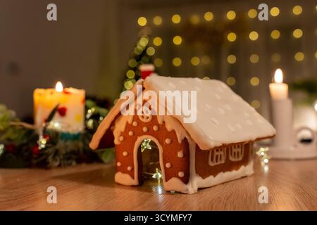 Schönes hausgemachtes Lebkuchenhaus mit weißer Glasur Dekoration und leuchtenden LED-Lichtern innen auf Holztisch mit Weihnachtskerzen und Bokeh Backgr Stockfoto