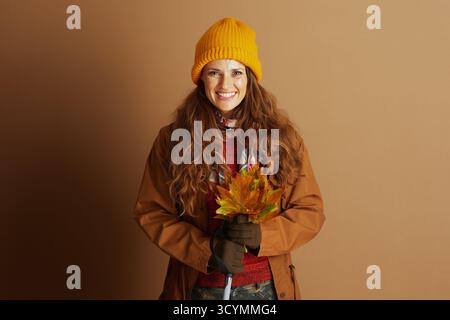 Eine lächelnde Frau, die in einer gelben Mütze, einer braunen Jacke und einem roten Pullover gekleidet ist, hält einen leuchtenden Blumenstrauß aus Herbstblättern. Das Bild fängt ein fröhliches und Wa ein Stockfoto