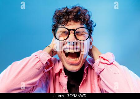 Nahaufnahme junger Mann in Brille, der vor Stress und Angst schreit, Kopfschmerzen und Burnout auf blauem Studiohintergrund ausdrückt. Stockfoto