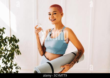 Junge Frau, die sich auf ein Training in einem hellen Raum mit einer Wasserflasche und Yogamatte vorbereitet Stockfoto